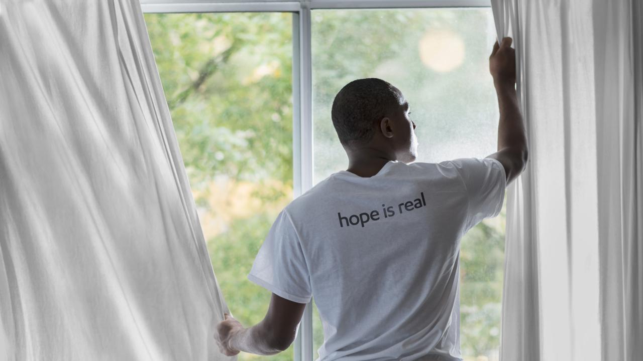 A man, drawing the curtain, wears a shirt that says Hope is Real